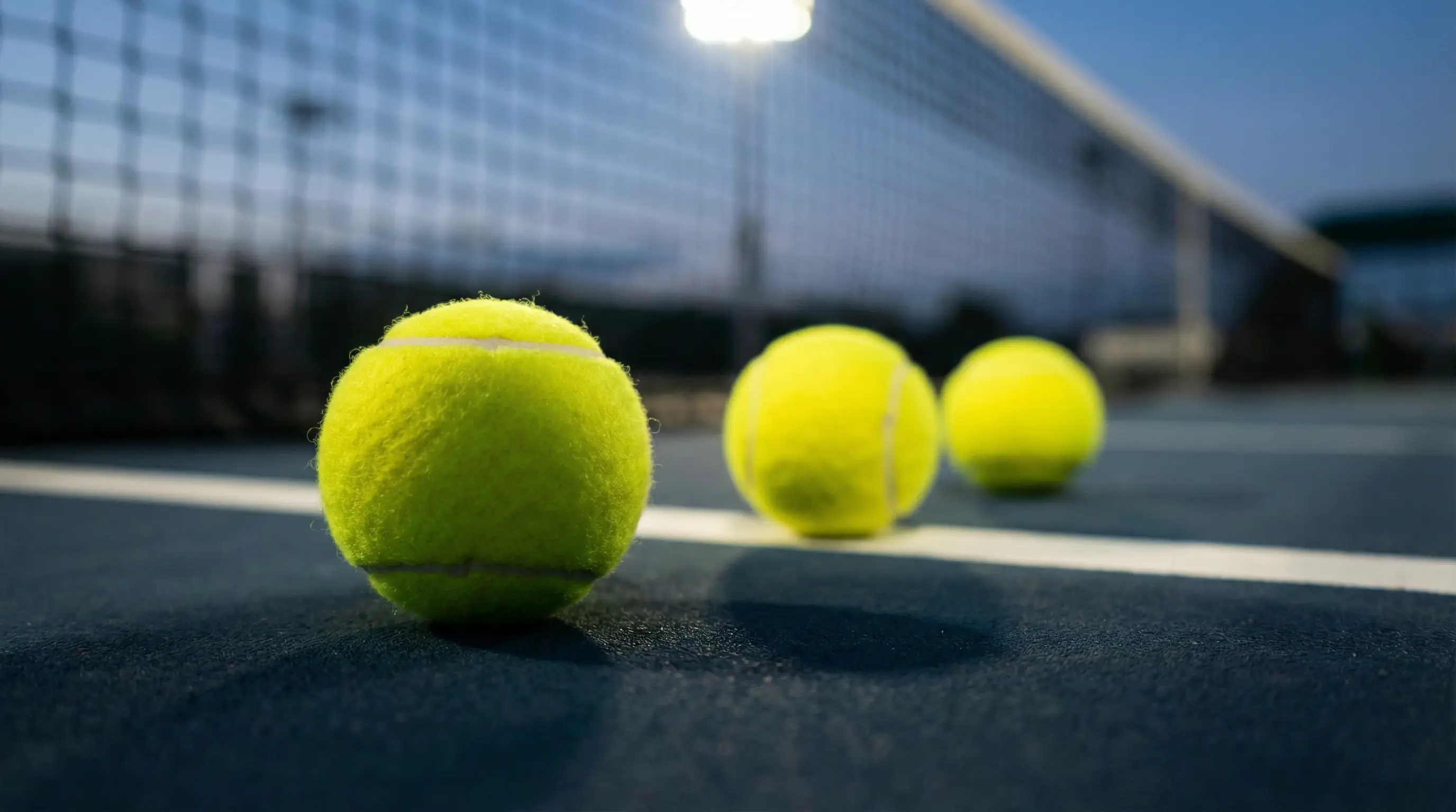 Plusieurs balles de tennis alignées sur un court en dur sous les projecteurs